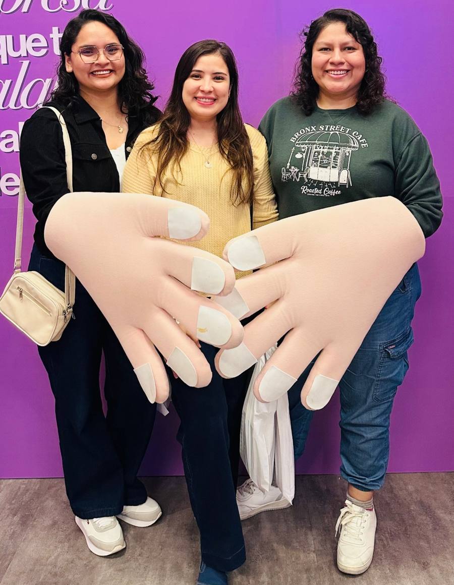 Tres mujeres sonríen juntas detrás de unas manos gigantes de tela que se entrelazan