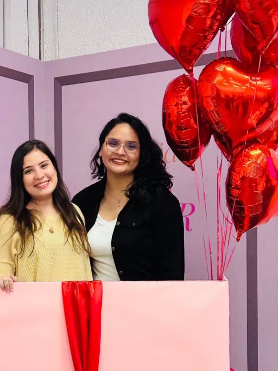 Dos mujeres sonríen dentro de una caja de regalo gigante, junto a globos de corazón